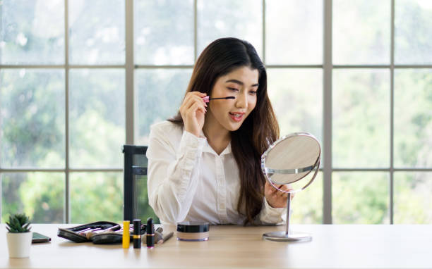 Woman applying mascara in front of a mirror with various makeup products on a table.